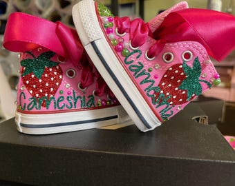 May include: Pink high-top trainers featuring a strawberry design and the name "Carneshia" in green glitter. The shoes have pink bows and decorative beads. The soles are white with a blue stripe.