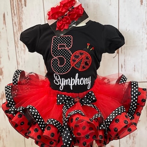 May include: A black t-shirt with a red and black ladybug design and the word "Symphony" in white script. The outfit includes a red tutu skirt with black polka dot ribbon trim and a matching bow. A set of red hair bows is also included.