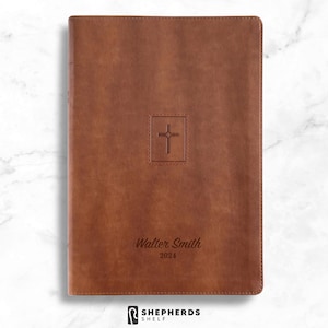 May include: A brown leather-bound book with a debossed cross emblem in a square frame. The name "Walter Smith" and the year "2024" are printed below the cross. The book has a simple, elegant design.
