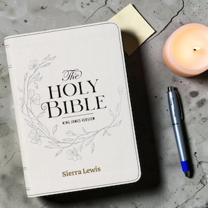 May include: A cream-colored Holy Bible King James Version with a floral wreath illustration on the cover. The text reads The Holy Bible, King James Version, and the name Sierra Lewis. The book features stitched edges and a matte finish.