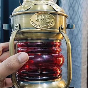 Könnte beinhalten: Antike Messinglaterne mit roter Glaslinse. Die Laterne hat einen Griff und der Text "LEEDS BURTON SOUTHAMPTON" ist sichtbar. Ein nautisches oder maritimes Thema.