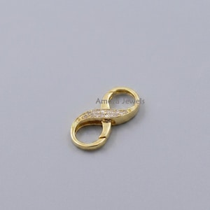 Op de afbeelding: Een goudkleurige oneindigheidssluiting met een rij kleine, heldere edelstenen. De sluiting heeft de vorm van een oneindigheidssymbool en heeft een scharniersluiting. De tekst "Amara Jewels" is zichtbaar.