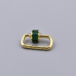 May include: A gold-colored jewelry clasp with a rectangular shape and a central green gemstone. The clasp has the text "Amora Jewels" on it. The clasp is set against a gray background.