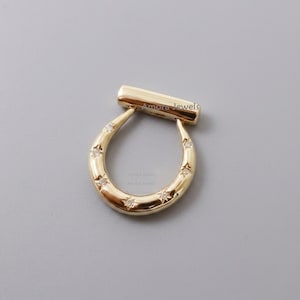Könnte beinhalten: Ein goldfarbener, hufeisenförmiger Anhänger mit kleinen, klaren Edelsteinen in Sternform. Der Anhänger hat oben einen rechteckigen Balken und der Text "Amora Jewels" ist sichtbar. Der Schmuck ist vor einem grauen Hintergrund platziert.