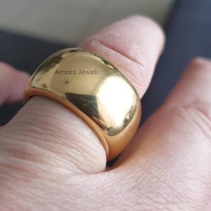 Peut inclure: Gros plan d'une bague dorée au design lisse et arrondi. La bague est portée au doigt et la surface reflète la lumière. Le texte "Amora Jewels" est visible sur la bague.