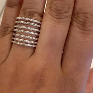 Könnte beinhalten: Ein silberner Ring mit mehreren Bändern, die jeweils mit kleinen, klaren Edelsteinen besetzt sind. Der Ring wird an einem Finger getragen und zeigt sein aufwendiges Design und seinen Glanz. Der Text "Amora Jewels" ist auf dem Ring zu sehen.