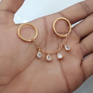 May include: Gold-coloured ring set connected by a delicate chain. The rings are linked with a chain that has five teardrop-shaped clear gemstones. The text "Amora Jewels" is visible on one of the rings.