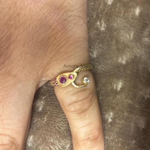 Könnte beinhalten: Ein goldfarbener Ring in Schlangenform, verziert mit rosa und klaren Edelsteinen. Der Ring befindet sich an einem Finger, mit dem Markennamen "Amora Jewels" sichtbar.