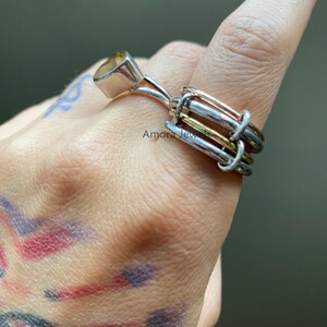 Könnte beinhalten: Ein Silberring mit einem gelben Edelstein und einem einzigartigen Mehrband-Design. Der Ring hat einen quadratisch geschliffenen gelben Stein in einem Silberband, mit zusätzlichen Bändern in Silber, Gold und Roségold. Der Ring ist an einem Finger.