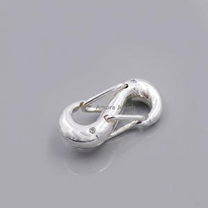 May include: A shiny silver-colored carabiner clasp with a curved design. The clasp has a spring-loaded gate and is marked with the text "Amora Jewels". The clasp is shown on a gray background.