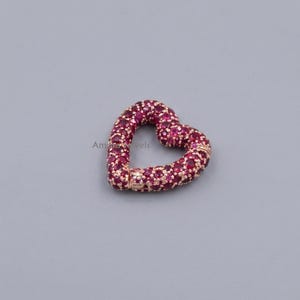 Puede incluir: Un cierre en forma de corazón, incrustado con numerosas piedras preciosas pequeñas y redondas de color rojo intenso. El cierre está engastado en un metal dorado, y el diseño es simétrico y elegante. El fondo es gris liso y apagado.