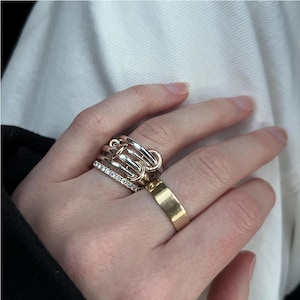 Könnte beinhalten: Nahaufnahme einer Hand mit mehreren Ringen. Ein breiter Goldring, ein Silberring mit kleinen, klaren Steinen und ein Ring mit einzigartigem Design.