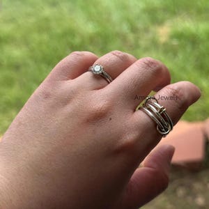 以下が含まれることがあります： 手には2つの指輪。1つは丸く透明な石が付いたシルバーの婚約指輪です。もう1つは、結び目のデザインと小さな金のアクセントが付いたシルバーの指輪です。背景はぼやけた緑色です。