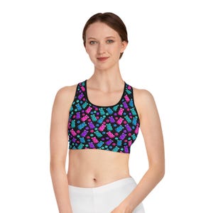 May include: A black sports bra with a colorful pattern of gummy bears in shades of blue, purple, and pink, along with matching polka dots. The bra has a scoop neck and racerback design.