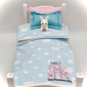 Pode incluir: Uma cama branca em miniatura com um cobertor azul e branco com padrão de nuvens e um conjunto de almofadas rosa e azul. Um pequeno coelho branco com um coração rosa está sentado no cobertor. O cobertor tem o texto "Cute PK Cute Persnickety Kids".