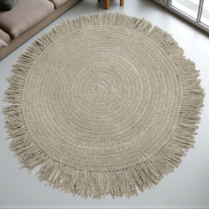 May include: A circular, beige jute rug with a fringed border. The rug features a woven, concentric circle pattern. It is placed on a light grey floor, with a sofa and window in the background.