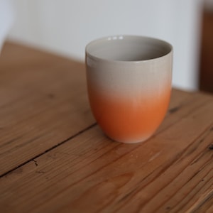Op de afbeelding: Een keramische beker met een gradiëntontwerp. De bovenste helft is lichtbeige, overgaand in een levendig oranje aan de onderkant. De beker staat op een houten oppervlak.