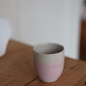Op de afbeelding: Een keramische beker met een tweekleurig ontwerp. De onderste helft is zachtroze, overgaand in lichtgrijs aan de bovenkant. De beker is cilindrisch met een afgeronde basis en staat op een houten oppervlak.