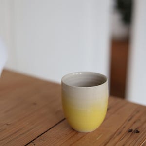 Op de afbeelding: Een keramische mok met een gele tot crèmekleurige gradiënt. De mok is cilindrisch met een afgeronde basis en staat op een houten oppervlak. De binnenkant van de mok is lichtgrijs.