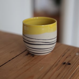Könnte beinhalten: Handgefertigte Keramiktasse mit gelbem Rand und cremefarbenem Körper. Der Körper der Tasse ist mit horizontalen schwarzen Streifen verziert. Die Tasse steht auf einer Holzoberfläche.