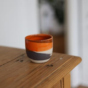 Könnte beinhalten: Eine kleine, handgefertigte Keramiktasse mit einem lebendigen, geschichteten Design. Die Tasse hat Bänder in Orange, Weiß und Dunkelgrau. Sie steht auf einer rustikalen Holzoberfläche und zeigt ihren einzigartigen, künstlerischen Stil. Die Tasse hat einen Durchmesser von etwa 7,6 cm.