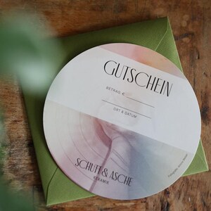 May include: A round gift certificate with the word "GUTSCHEIN" in black, resting on a green envelope. The certificate has a watercolor design in pastel colors. The text "SCHUFF & ASCHE" is also visible.