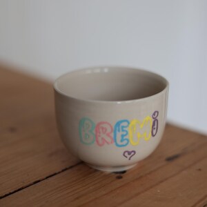 Könnte beinhalten: Beige Keramikschale mit dem Wort "BREMI" in bunten Buchstaben. Ein kleines lila Herz befindet sich unter dem Wort. Die Schale steht auf einer Holzoberfläche.