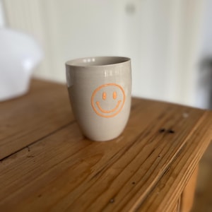 Peut inclure: Un mug en céramique beige avec un motif de visage souriant orange. Le mug a une forme cylindrique et repose sur une surface en bois. Le visage souriant est centré sur le devant du mug.