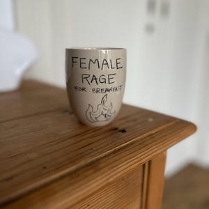 Może przedstawiać: Beżowy ceramiczny kubek z napisem "FEMALE RAGE FOR BREAKFAST" czarną, odręczną czcionką. Poniżej znajduje się mała ilustracja płomieni i bananów. Kubek stoi na drewnianym stole.