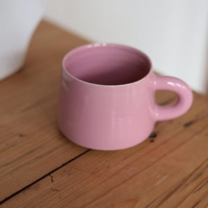 Könnte beinhalten: Eine rosa Keramiktasse mit rundem Körper und gebogenem Henkel. Die Tasse hat eine einfarbige, weiche rosa Farbe und steht auf einer Holzoberfläche. Die Tasse ist leer und scheint sauber zu sein.