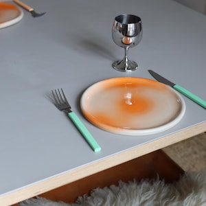 Könnte beinhalten: Ein gedeckter Tisch mit Teller, Besteck und Weinglas. Der Teller hat ein orange-weißes Farbverlauf-Design. Gabel und Messer haben mintgrüne Griffe. Das Weinglas ist aus Edelstahl.