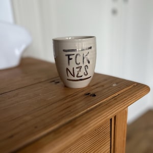 Może przedstawiać: Beżowy ceramiczny kubek z czarnym napisem "FCK NZS". Kubek jest cylindryczny i stoi na drewnianej powierzchni. Tło to rozmyta biała ściana.