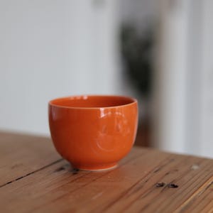 Puede incluir: Un pequeño cuenco de cerámica naranja con un diseño de carita sonriente. El cuenco está sobre una superficie de madera. El cuenco mide aproximadamente 7,6 cm de diámetro y 5 cm de alto. El fondo está borroso.