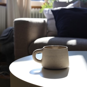 Könnte beinhalten: Eine beige Keramik-Tasse mit rundem Henkel steht auf einem weißen Tisch. Die Tasse hat eine strukturierte Oberfläche und steht im Sonnenlicht. Der Hintergrund umfasst ein graues Sofa und ein Fenster mit Vorhängen.
