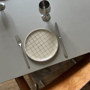 Op de afbeelding: Een tafelsetting met een crèmekleurige bord met een zwart rasterpatroon, een roestvrijstalen mes, vork en beker. Extra borden en bestek zijn zichtbaar op de achtergrond. De tafel is grijs.
