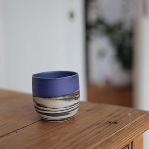 Könnte beinhalten: Eine handgefertigte Keramiktasse mit blauem Rand und einem Design aus horizontalen Streifen in Weiß-, Braun- und Schwarztönen. Die Tasse steht auf einer Holzoberfläche.