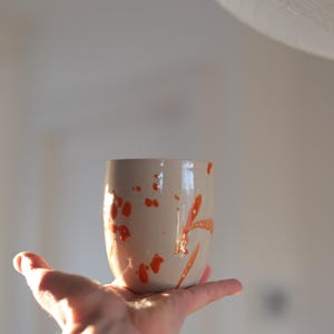 Op de afbeelding: Een beige keramische beker met levendige oranje verfspatten. De beker wordt in een hand gehouden en toont zijn unieke, artistieke ontwerp. Een handgemaakt item, perfect voor koffie of thee.
