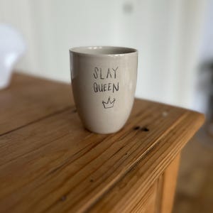 Könnte beinhalten: Eine beige Keramiktasse mit den Worten "SLAY QUEEN" und einer Kronenillustration in Schwarz. Die Tasse ist zylindrisch und steht auf einer Holzoberfläche. Der Hintergrund ist unscharf.
