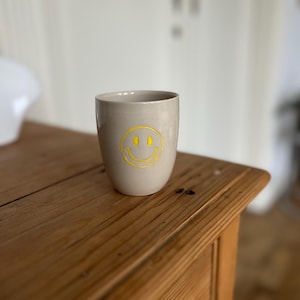 Könnte beinhalten: Eine beige Keramiktasse mit einem gelben Smiley-Gesicht. Die Tasse hat eine zylindrische Form und steht auf einer Holzoberfläche. Das Smiley-Gesicht ist mittig auf der Vorderseite angebracht.