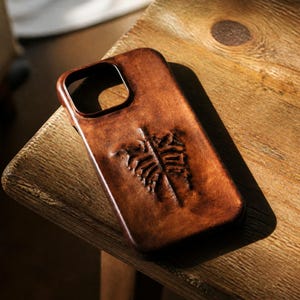 Könnte beinhalten: Eine braune Handyhülle aus Leder mit einem rechteckigen Kameraausschnitt und einem geprägten Farnblatt-Design auf der Rückseite. Die Oberfläche hat ein abgenutztes, strukturiertes Finish mit dunkelbraunen Schattierungen und Glanz.