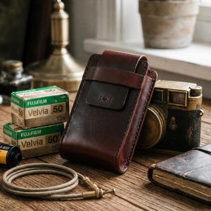 May include: A dark brown leather pouch with a flap closure and a strap, featuring the embossed letters J and T with a heart symbol in between. The pouch is positioned on a wooden surface alongside two boxes of Fujifilm Velvia 50 film and a camera.