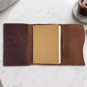May include: A dark brown leather notebook cover featuring multiple interior card slots and a vertical pocket on the left side, with a tan paper notebook inserted into the center sleeve and a matching leather flap on the right side.