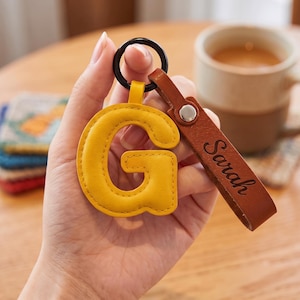 Puede incluir: Un llavero de cuero amarillo con forma de letra G unido a una anilla de metal negro y una tira de cuero marrón con el nombre Sarah grabado en letra cursiva negra, asegurado con un remache de metal plateado.