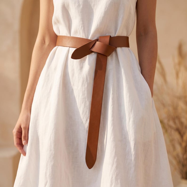 May include: A brown leather belt tied in a knot, worn over a white dress. The belt's color contrasts with the dress, creating a visual focal point. The dress has a simple design, and the belt adds a stylish accent.
