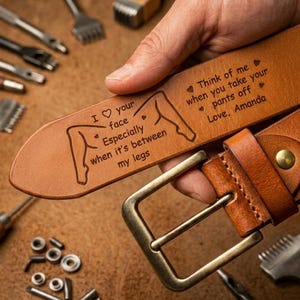 May include: A tan leather belt with a rectangular brass buckle. The belt is engraved with the text "I ♡ your face Especially when it's between my legs" and "Think of me when you take your pants off Love, Amanda."