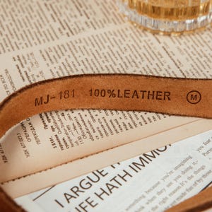 May include: Close-up of a brown leather belt with the text "MJ-181 100% LEATHER" stamped on it. The belt is lying on top of a newspaper. A small "M" is stamped on the belt.