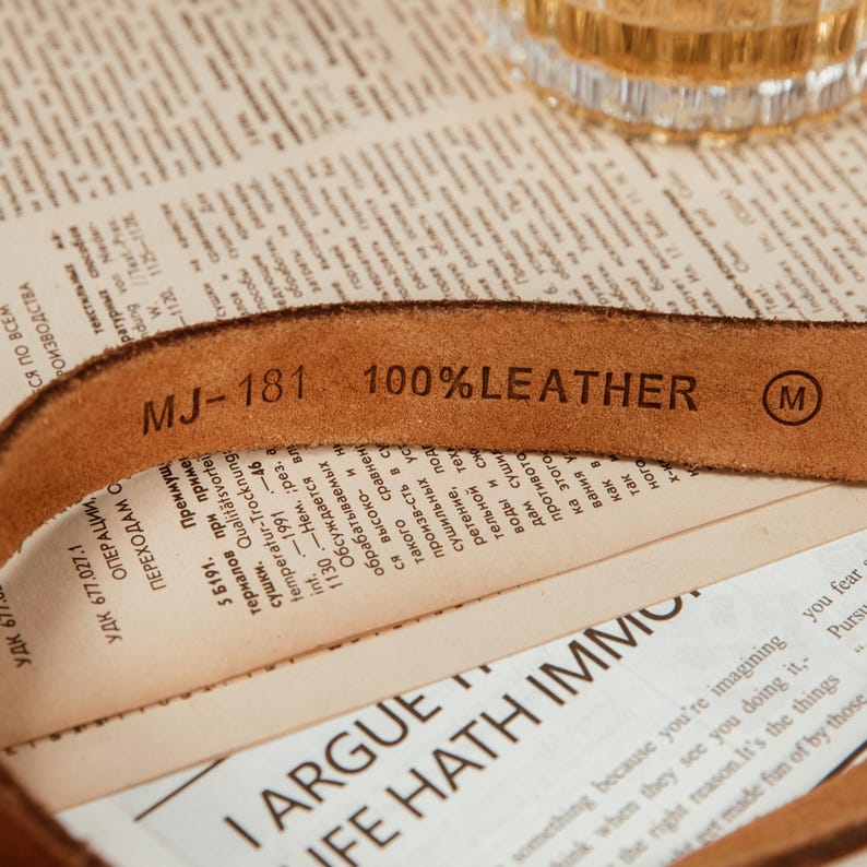 May include: Close-up of a brown leather belt with the text "MJ-181 100% LEATHER" stamped on it. The belt is lying on top of a newspaper. A small "M" is stamped on the belt.