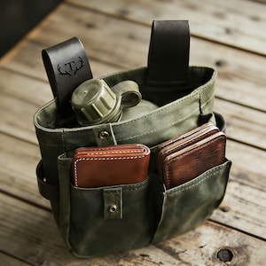 Pode incluir: Uma bolsa utilitária de lona verde-oliva com duas alças de cinto de couro marrom escuro, uma delas gravada com uma letra T e um logotipo de chifres. A bolsa contém um cantil de plástico verde e duas carteiras de couro marrom nos bolsos.