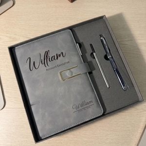 Puede incluir: Un cuaderno gris con un cierre rectangular dorado y un bolígrafo a juego en una caja de regalo gris. La portada presenta el nombre William, el título Account Executive y el nombre William con la fecha 14 de junio de 2026 en texto negro.