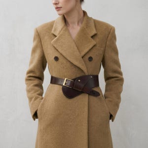 May include: A tan double-breasted coat with a dark brown leather belt. The belt has a gold-coloured buckle and wraps around the waist. The coat has two rows of buttons and pockets.